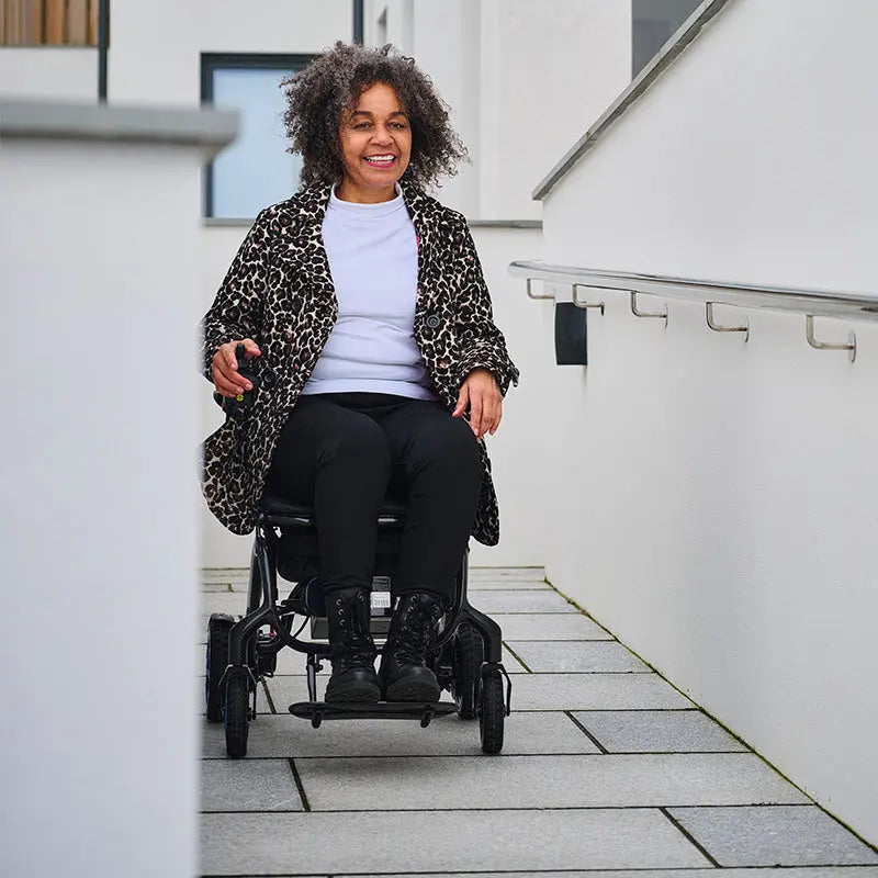 Person in a leopard print coat using a Quickie Q50 R Carbon Folding Powerchair on a sidewalk.