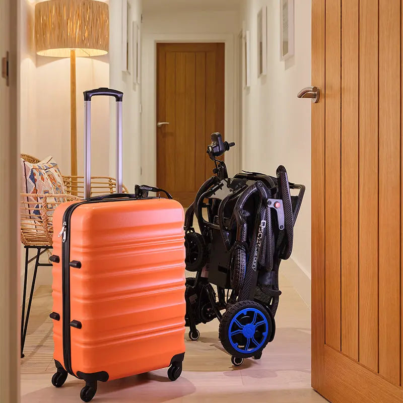 Orange suitcase and folded Quickie Q50 R Carbon Folding Powerchair in a room with wooden door and lamp.