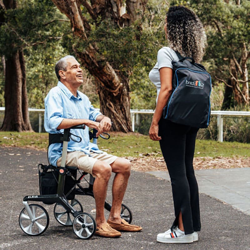 A man in an aspire ninja carbon fibre lightweight seat walker is being assisted by a woman with a backpack outdoors.
