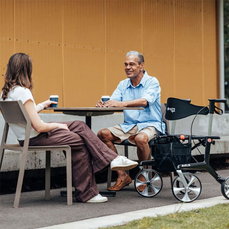 Two people sitting at a table outdoors with a person using the aspire ninja carbon fibre lightweight seat walker.
