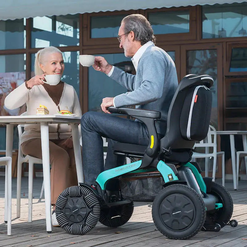Man using Robooter E60 Pro All Terrain Electric Folding Powerchair outdoors with a woman sitting at a table.