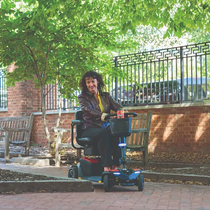 Woman using a Pride Jazzy Zero Turn Travel Scooter in an outdoor setting with trees and a building in the background.