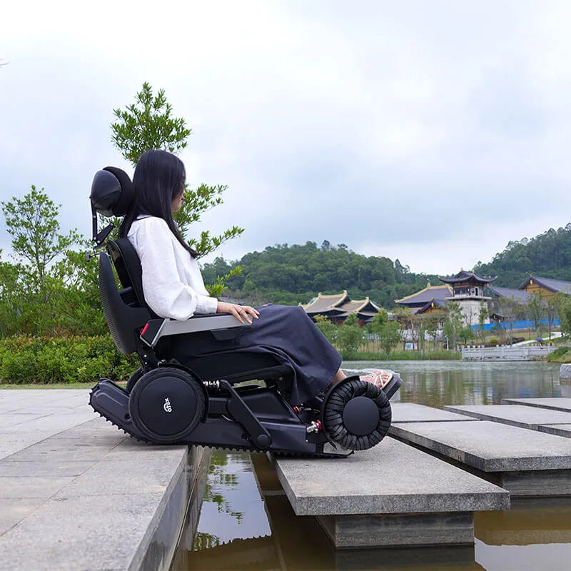 Person using a Xsto X12 All-Terrain Mobility Robot Powered Wheelchair
 by a scenic lake with traditional architecture.