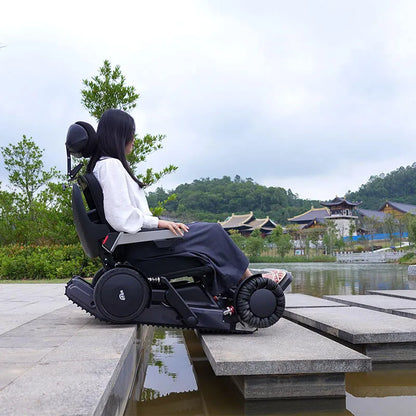 Person using a Xsto X12 All-Terrain Mobility Robot Powered Wheelchair
 by a scenic lake with traditional architecture.