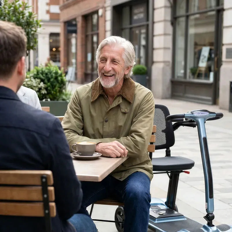Man sitting at an outdoor cafe table with a coffee, smiling and engaged in conversation. AeroMg Lightweight Folding Mobility Scooter in the background