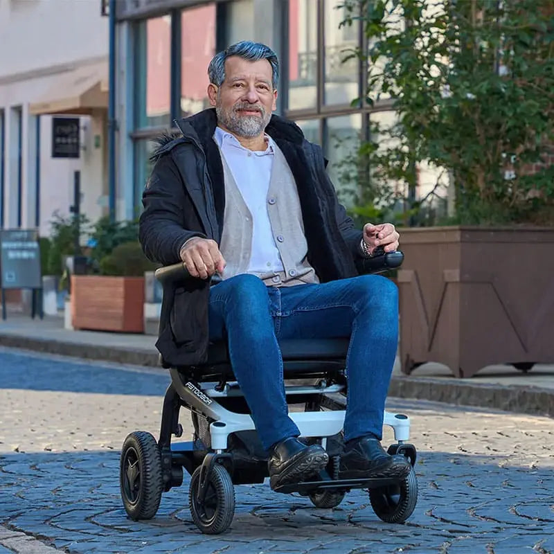 Man using a Robooter E80 Ultra Carbon Fiber Lightweight Folding Powerchair on a sidewalk with buildings and plants in the background
